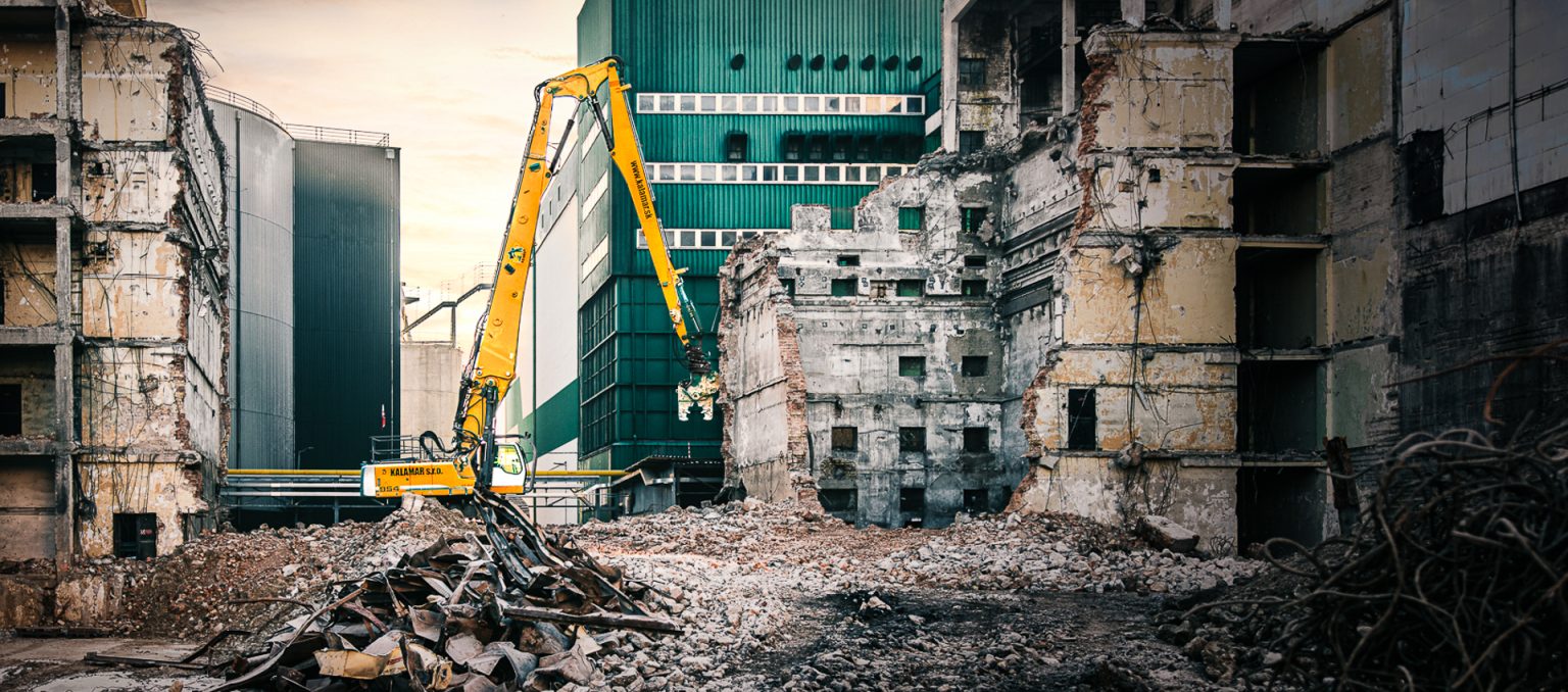 Pásové rýpadlo Liebherr R 954 C spoločnosti Kalamár s.r.o. s dlhým ramenom a búracou nožnicou v priemyselnom areáli pri demolovanej tehlovej budove. Demolačné práce.