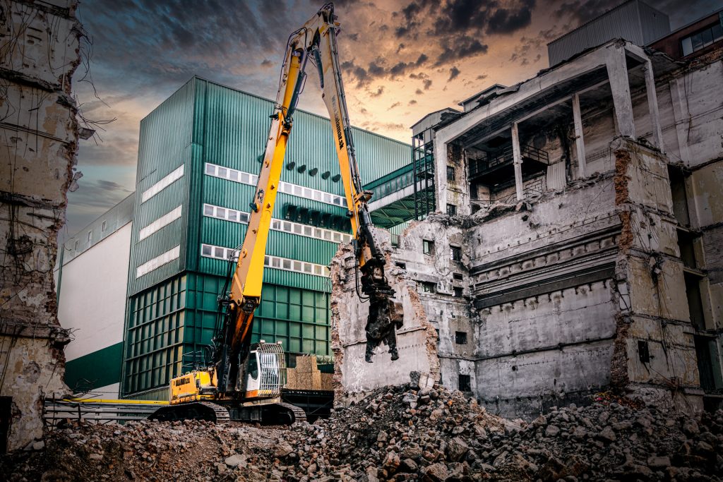 Pásové rýpadlo Liebherr R 954 C spoločnosti Kalamár s.r.o. s dlhým ramenom a búracou nožnicou v priemyselnom areáli pri demolovanej tehlovej budove. Demolačné práce.