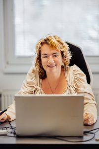 Žena v béžovej košeli sediaca za pracovným stolom pred oknom v kancelárii. Na stole má notebook.